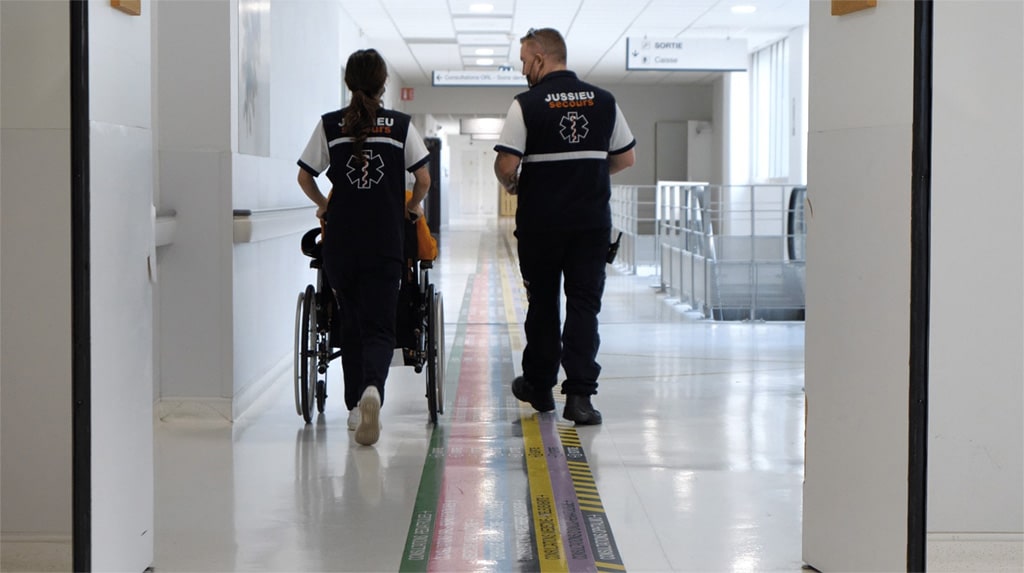 Photo d’illustration : dans un couloir du GHT, deux ambulancier·ère poussent un fauteuil roulant